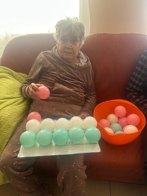 Seniorka na kanapie sortuje kolorowe piłeczki, układając je rzędami obok pomarańczowej miski w CuratioW w Rajczy