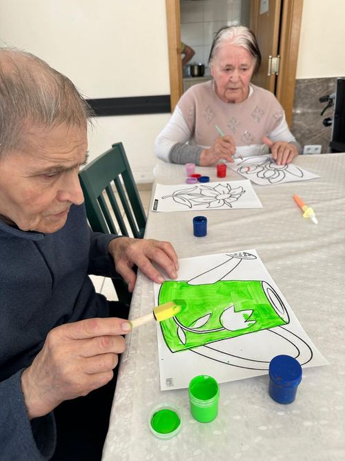 Dwoje seniorów przy stole maluje farbami kolorowanki z motywami kwiatów podczas warsztatów plastycznych w CuratioW