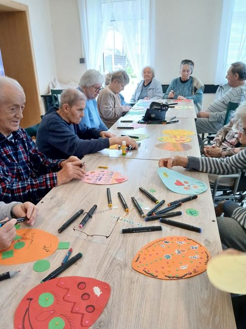Grupa seniorów przy długim stole ozdabia papierowe pisanki mazakami podczas warsztatów wielkanocnych w CuratioW