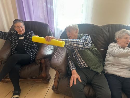 Seniorzy w fotelach ćwiczą sprawność rąk przesuwając żółtą piankę po napiętym sznurku podczas zabawy ruchowej w CuratioW