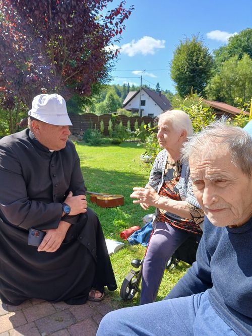 Ksiądz rozmawia z seniorami w ogrodzie CuratioW w Rajczy w słoneczny letni dzień