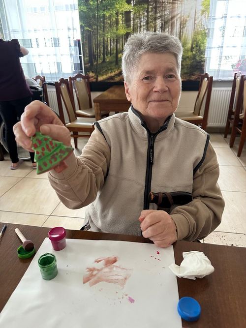 Seniorka podczas warsztatów malarstwa prezentuje pomalowaną na zielono drewnianą choinkę przy stole z farbkami w CuratioW