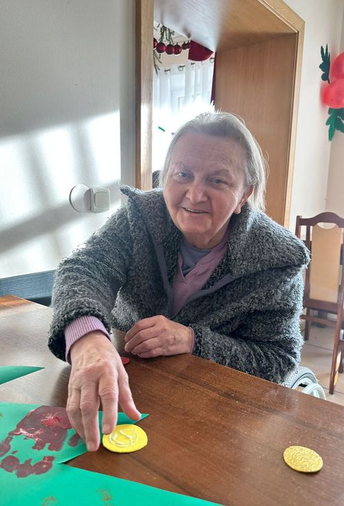 Seniorka w szarym swetrze nakleja żółte brokatowe kółka na zieloną kartkę podczas zajęć rękodzielniczych w Rajczy