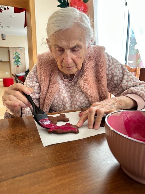 Seniorka pędzlem dekoruje pierniczki różowym lukrem przy stole, obok miska z polewą, podczas warsztatów kulinarnych w CuratioW