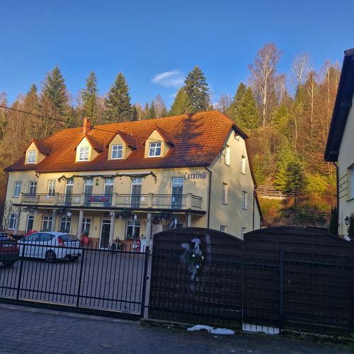 Front CuratioW w pełnym lecie, żółta elewacja, balkon i czerwone krzesła przy wejściu, otoczony bujną zielenią i świerkami