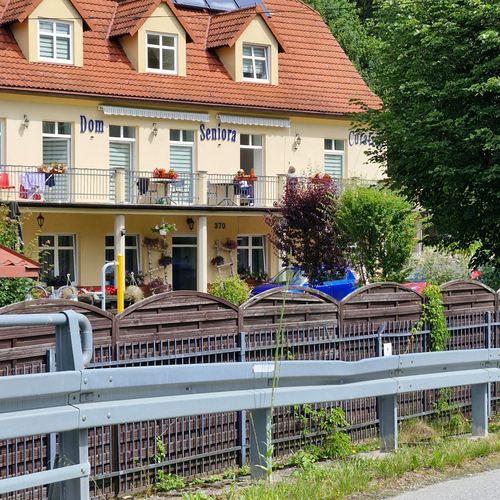 Frontowa fasada CuratioW z napisem Dom Seniora, kwiaty na balkonach i drewniany płot latem